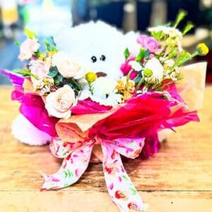 A white teddy bear surrounded by colorful flowers and pink wrapping on a wooden table.